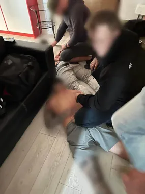 W pomieszczeniu mieszkalnym kilka osób siedzi na podłodze przy łóżku, jedna z nich trzyma nogę innej osoby.
