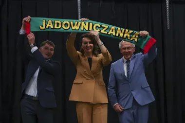 Trzy osoby w eleganckich garniturach unoszą zielony szalik z żółtym napisem JADOWNICZANKA na tle ciemnej kurtyny.