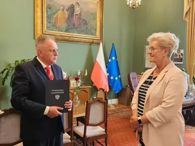 Mężczyzna w garniturze trzyma teczkę z napisem „Wojewoda Małopolski” podczas spotkania z kobietą w eleganckim gabinecie z flagami Polski i UE.